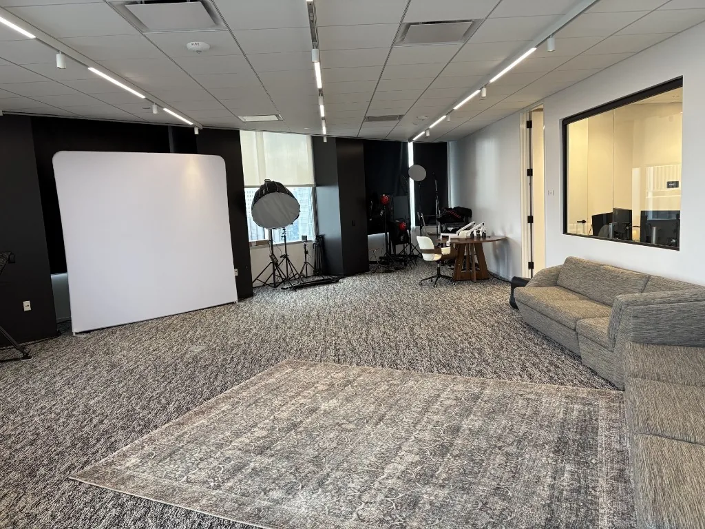Existing NRG studio showing a grey sectional sofa, an ornate area rug, a white fabric backdrop against a black wall, lighting stands near the windows, and an interior window into an adjacent control room.