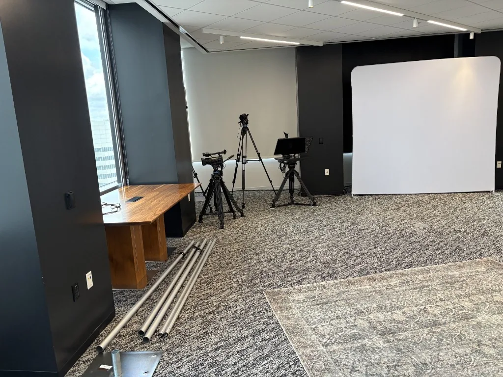 Existing NRG studio production area with camera tripods, a teleprompter, a portable white backdrop, a wooden desk against a structural pillar, and metal grip poles on grey office carpet.
