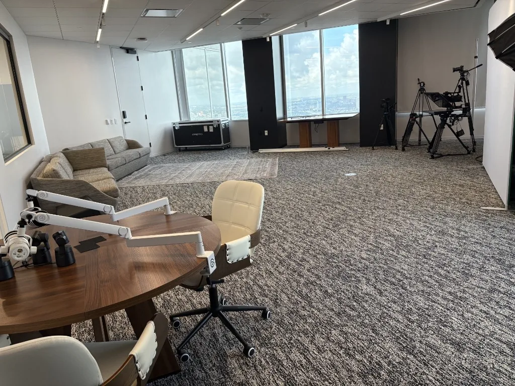Wide view of the existing NRG video production studio: an open high-rise room with a round podcast table, boom microphones, a grey lounge sofa, equipment cases, camera tripods, and floor-to-ceiling windows.